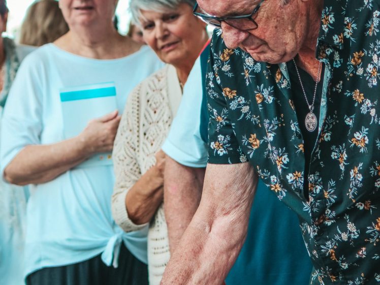 Image of An elderly man in a floral shirt serves himself food from a table while several older women stand in queue behind him, waiting their turn. The setting appears to be a casual indoor gathering.