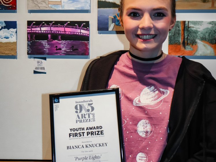 Image of A smiling young person holding a framed certificate for the Youth Award First Prize in front of a wall displaying various small artworks. The certificate reads Bianca Knuckey, Purple Lights.