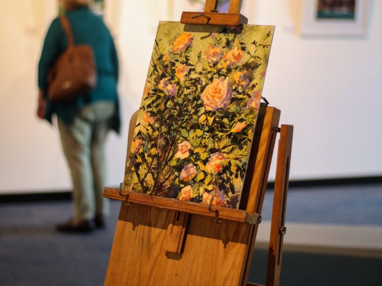 Image of An easel with a colourful painting of flowers is in focus in the foreground, while a person with a brown bag looks at framed artworks on a gallery wall in the blurred background.