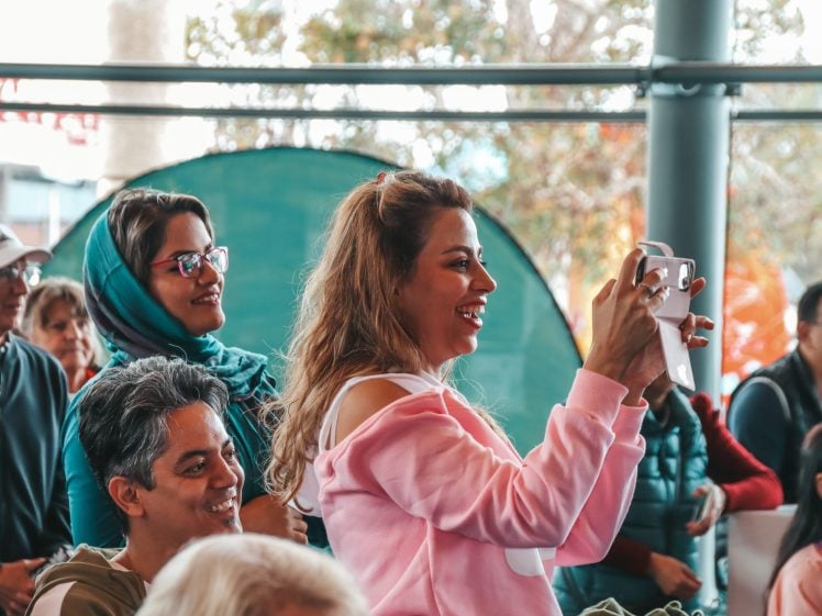 Image of A woman in a pink hoodie smiles and takes a photo with her mobile in a lively, diverse indoor crowd. Other people around her are also watching something with interest.