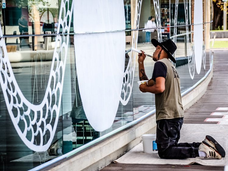 Image of A person kneels on the pavement, painting a white, scale-like pattern on large glass windows. They wear a cap, mask, and casual clothes, holding a paintbrush in one hand and a palette in the other.