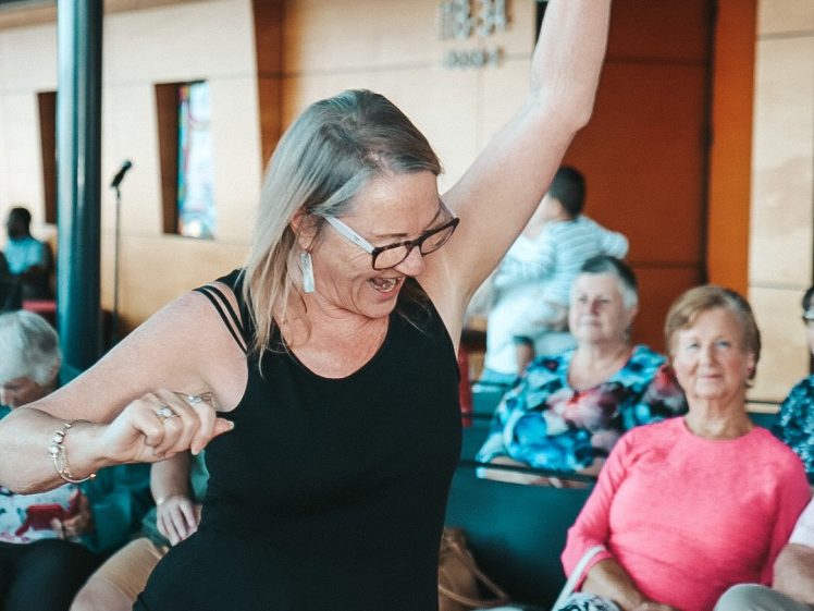 Image of A woman in a black vest top and pink skirt dances joyfully with one arm raised, while several older adults seated in the background watch and smile. The setting appears to be indoors, possibly at a community event.