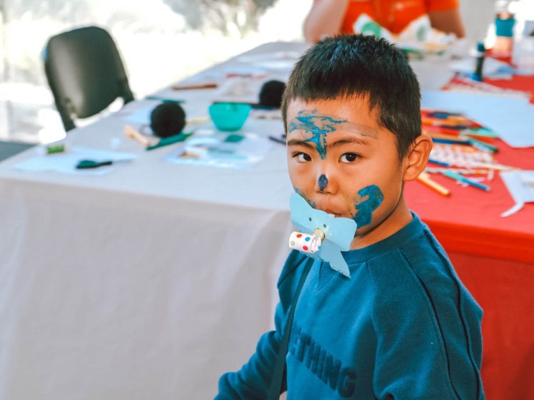 Image of A young boy with blue paint on his face and a paper craft in his mouth stands near a table covered with art supplies and colouring materials.
