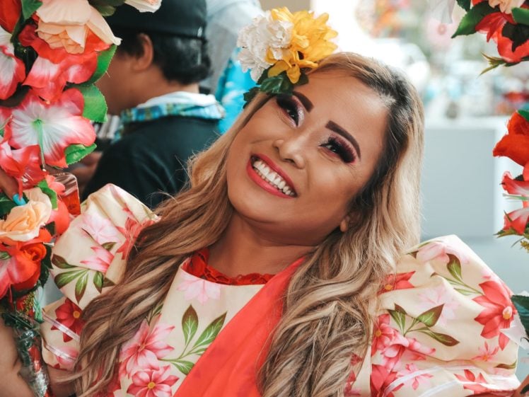 Image of A smiling person with long blonde hair wears a floral dress and vibrant make-up, holding colourful flower garlands. They have a yellow and white flower in their hair, celebrating at an outdoor event. Other people are in the background.