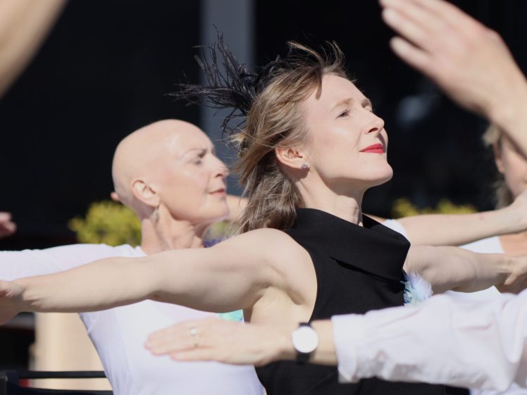 Image of Two women stand with arms outstretched, eyes closed, and faces lifted as they enjoy the sun. Others’ arms are visible in the foreground. One woman has short hair and the other is bald; both appear serene and joyful.