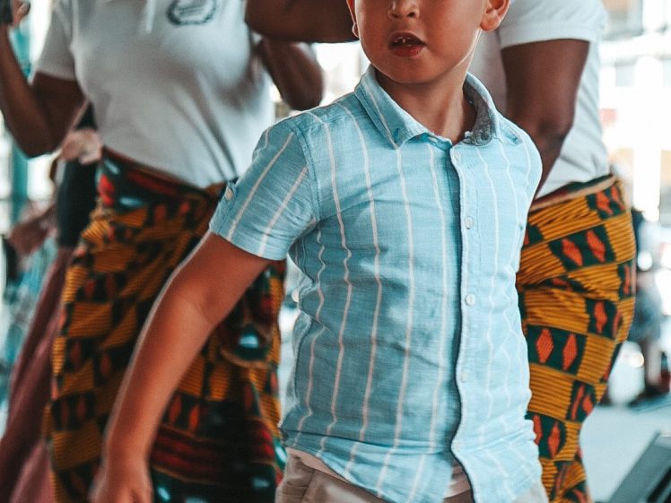 Image of A young boy in a blue striped shirt dances in front of two women in patterned skirts and white shirts, who are also dancing and wearing matching colourful headbands.