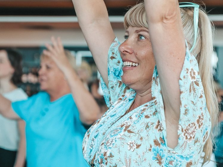 Image of A smiling woman with blonde hair and a floral shirt raises her arms enthusiastically in a group setting, with other people in the background also participating and appearing engaged.