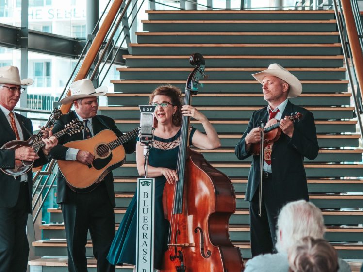 Image of A bluegrass band performs indoors in front of a staircase. The group includes three men in suits and cowboy hats playing guitar, mandolin, and fiddle, and a woman in a dress playing a double bass. An audience watches them.