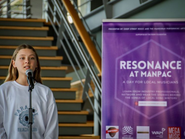 Image of A young woman sings into a microphone next to a large purple banner that reads “Resonance at MANPAC—A Day for Local Musicians.” A staircase and railing are visible in the background.