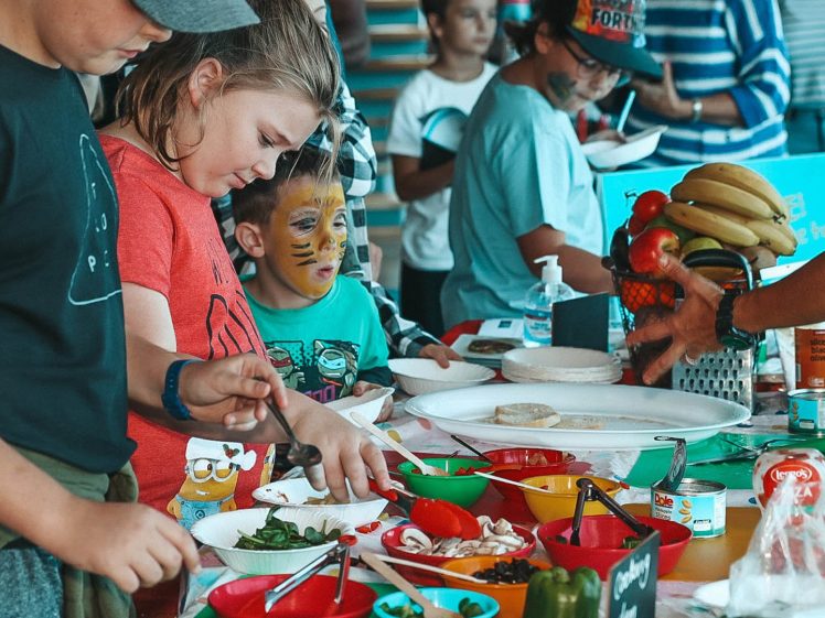Image of Children with painted faces and adults gather around a colourful table filled with bowls of vegetables, fruit, and other food items, preparing or serving food at what appears to be a festive indoor event.
