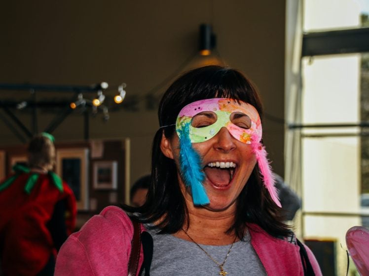 Image of A woman wearing a colourful, feathered mask and a pink jacket laughs joyfully indoors, with blurred people and a window in the background.