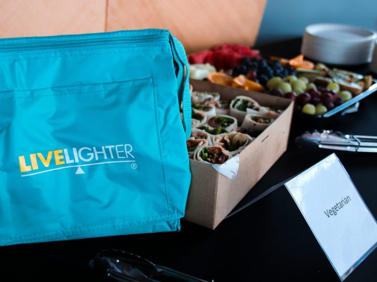 Image of A turquoise LIVELIGHTER insulated lunch bag sits beside a tray of assorted vegetarian wraps and fruit, with plates, tongs, and a Vegetarian sign on a black tablecloth.