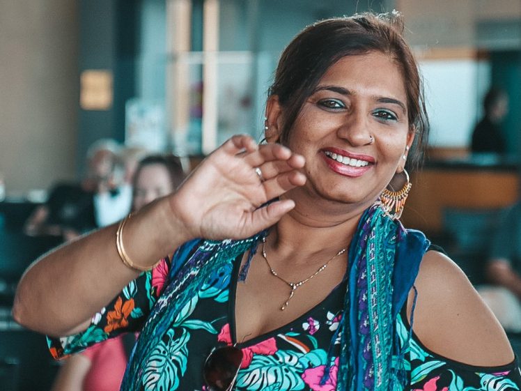 Image of A woman in a colourful floral outfit smiles and waves at the camera inside what appears to be an airport or waiting area, with people and a box office sign in the background.