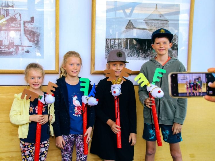 Image of Four children stand in a row, smiling and holding reindeer hobby horses. One person is taking a photo of them with a mobile phone. Framed black-and-white pictures hang on the wall behind them.