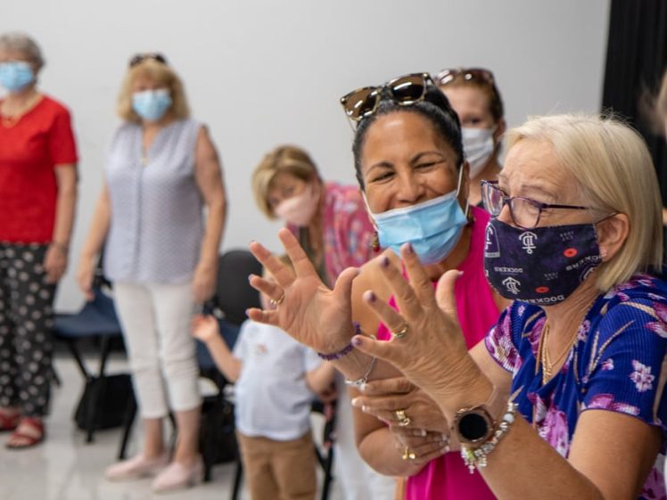 Image of A group of people wearing face masks stand indoors, some smiling and gesturing with their hands, suggesting a lively and friendly conversation or activity.