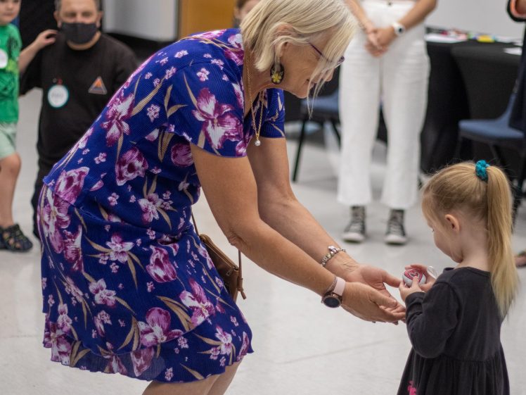 Image of An older woman in a blue floral dress bends down to hand a small object to a young girl with a ponytail. Several people, including children and adults, are visible in the background of a brightly lit room.