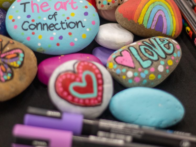 Image of Colourful painted rocks with positive words like The art of connection, JOY, and LOVE, along with hearts, flowers, and a rainbow, are arranged on a table next to several coloured markers.