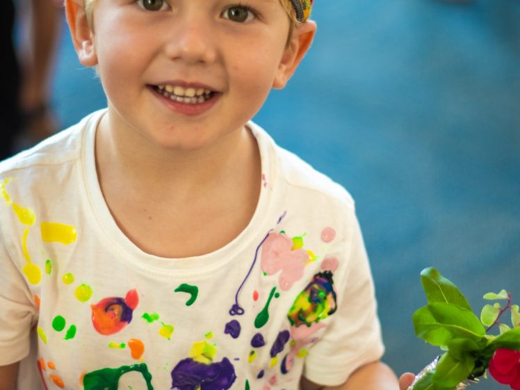 Image of A smiling young child wearing a colourful striped hat and a white shirt with bright paint splatters holds a small bunch of flowers. The background is blurred with people and blue flooring visible.