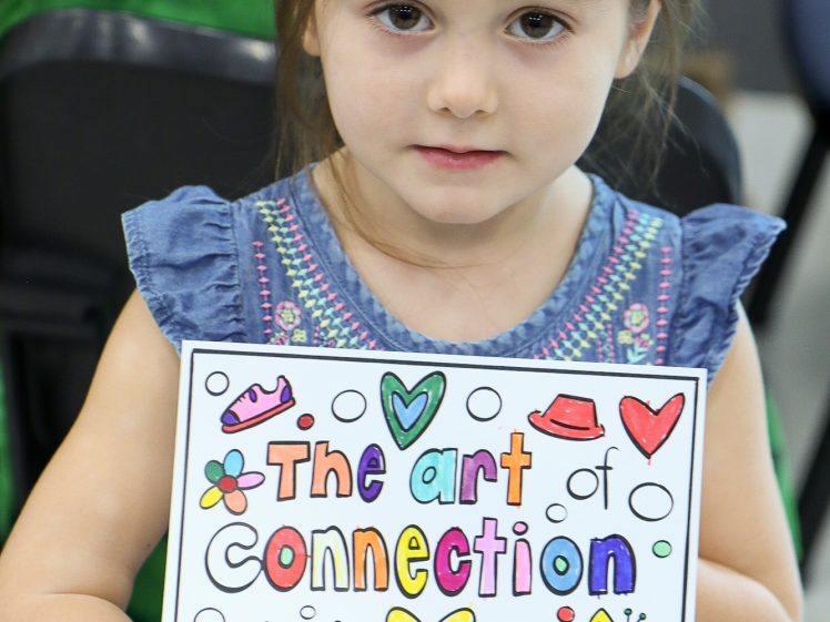 Image of A young girl in a blue dress holds up a colourful sign that reads The art of connection, decorated with hearts, stars, a rainbow, a cupcake, and other playful drawings.
