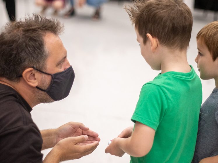 Image of A man wearing a face mask crouches and speaks to two young boys standing side by side. The man is gesturing with his hands, and the boys are facing him, listening attentively. The background is slightly blurred.