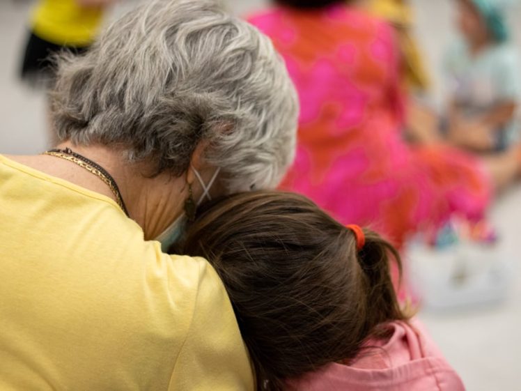 Image of An older woman with grey hair and a young girl sit closely together, with the girl's head resting on the woman's shoulder. Both are facing away, and there are blurred people and bright colours in the background.