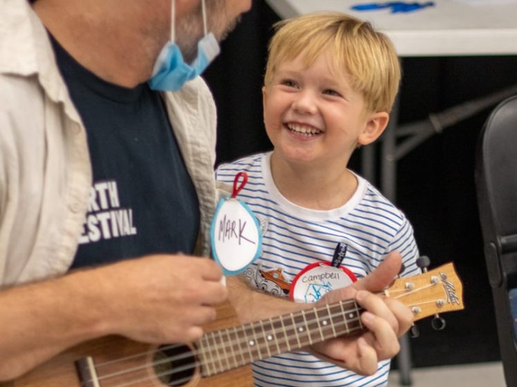 Image of An adult wearing a hat and a child sit together; the adult plays a ukulele while the child smiles joyfully. Both have name tags, and the scene appears cheerful and interactive.