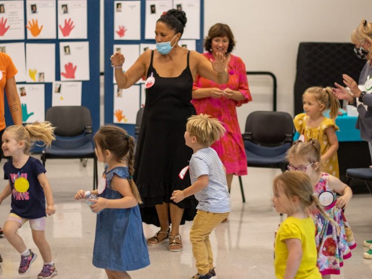 Image of A group of young children dance and play in a classroom with several adults supervising. The kids appear joyful and energetic. One adult wears a mask, and colourful artwork is displayed on the wall behind them.