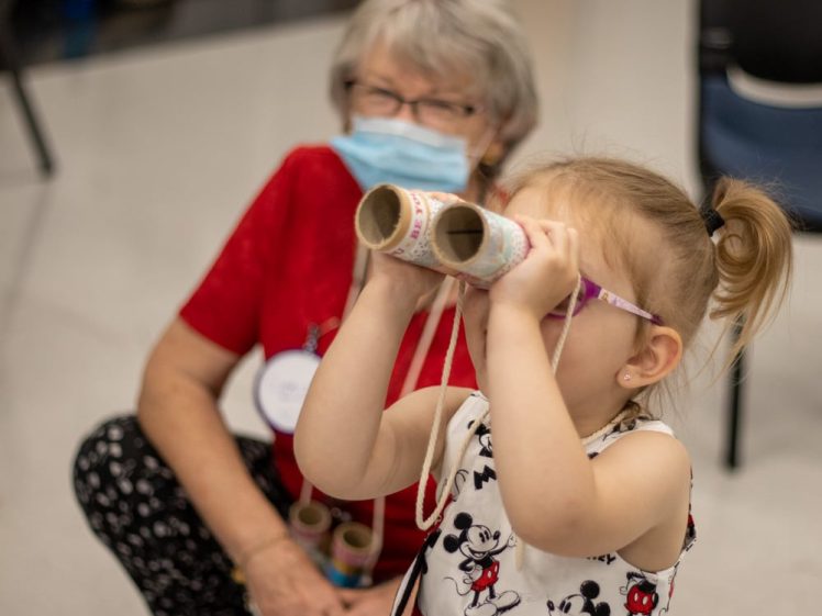 Image of A young girl wearing glasses looks through homemade binoculars made from decorated cardboard tubes, while an older woman in a mask and red shirt sits nearby, watching her.