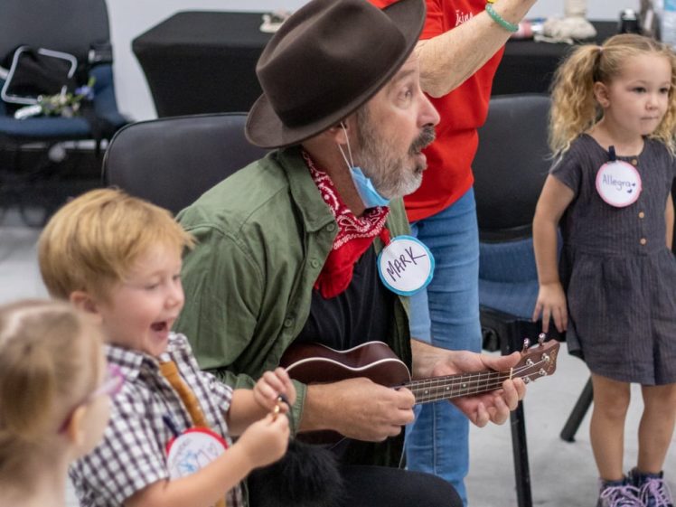 Image of A man wearing a hat and red scarf strums a ukulele while kneeling among smiling young children in a classroom setting. An adult in a red shirt stands behind them, engaging with the children.