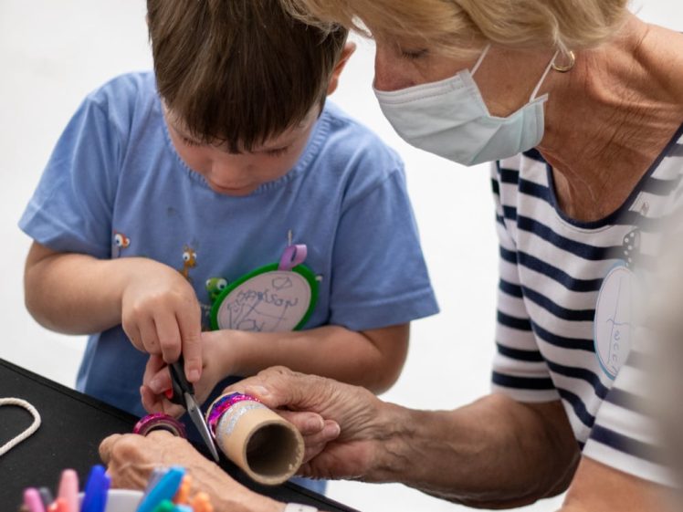 Image of A child and an older woman wearing a mask work together on a craft project, cutting a decorated cardboard tube. Colourful markers and string are nearby on the table.