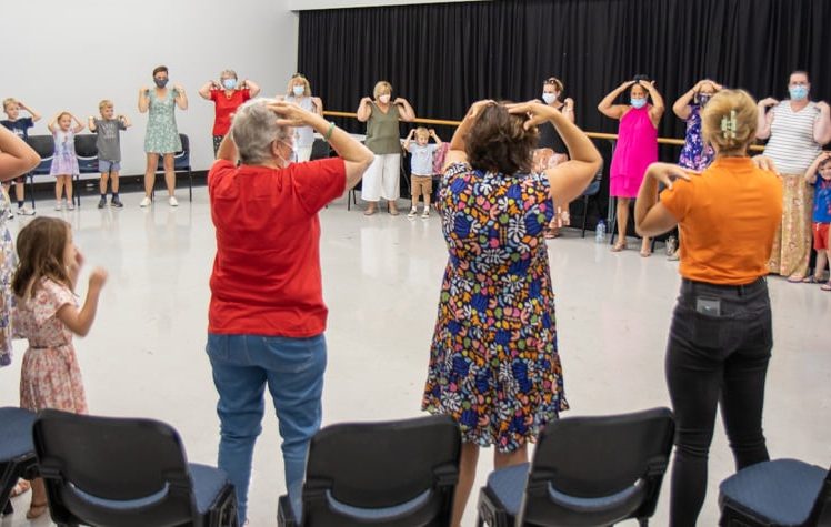 Image of A group of people, including adults and children, stand in a circle in a brightly lit room, mimicking hand movements above their heads during an activity or workshop. Some participants are wearing face masks.
