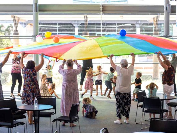Image of Adults and children play indoors with a large, colourful parachute. Some children are underneath while adults and a few kids hold the edges, lifting it up. Chairs and tables are scattered around the bright, spacious room.