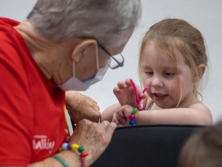 Image of An elderly woman wearing a mask and red shirt helps a young girl with a craft project using colourful beads and pipe cleaners. They are both focused on the activity, sitting closely together.