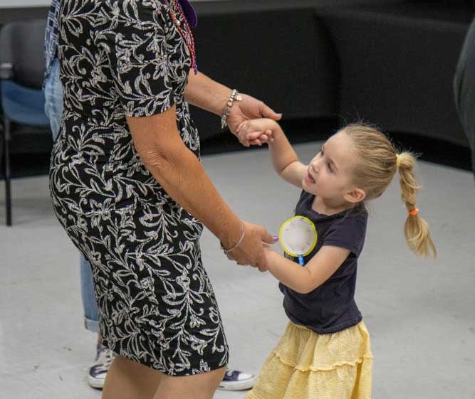 Image of An older woman in a black and white dress dances with a smiling young girl in a yellow skirt and dark top. They hold hands and make eye contact in a bright indoor room.