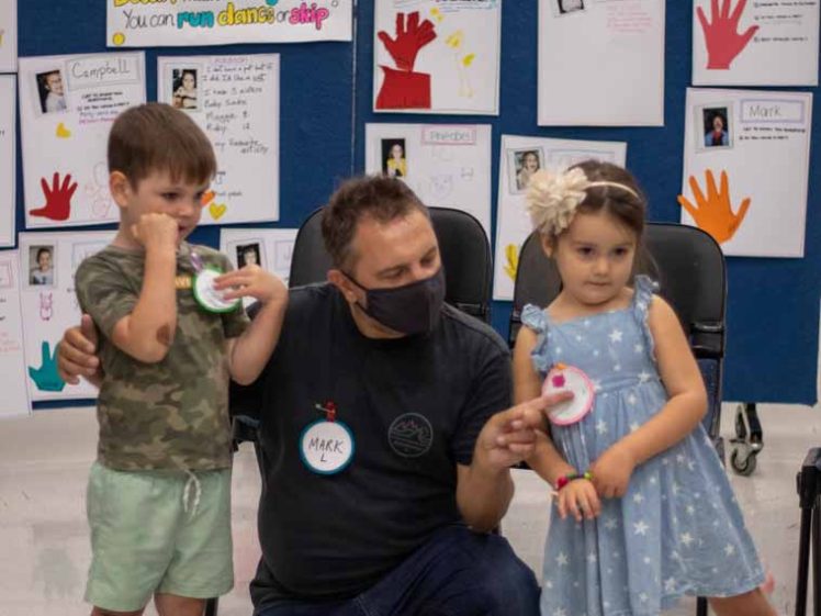 Image of A man wearing a mask kneels between a young boy and girl in a classroom. Colourful posters and children’s artwork are displayed on the wall behind them. The children wear name tags and the girl holds a ribbon.