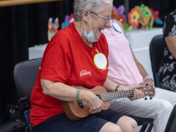 Image of An older woman wearing a red shirt and glasses sits on a chair, smiling and playing a ukulele. She has a name tag and a mask pulled under her chin. Colourful paper flowers are displayed in the background.