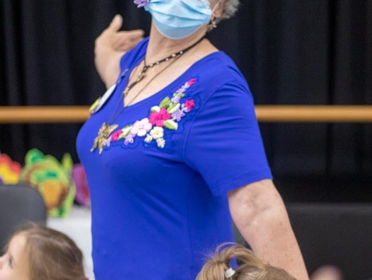 Image of An older woman wearing a blue shirt with colourful flower embroidery and a face mask poses playfully. She has flowers and a butterfly hair accessory. Two young children are nearby, and the background is blurred.