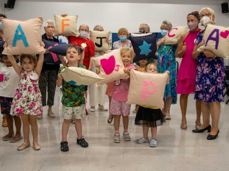 Image of A group of children and adults stand indoors, each holding handmade cushions decorated with colourful letters and shapes. Some people wear masks, and everyone appears happy and engaged in the activity.
