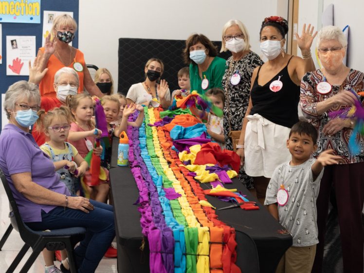 Image of A group of adults and children, many wearing masks, gather around a table covered with colourful fabric strips. They are smiling, waving, and appear to be working on a collaborative art project indoors.