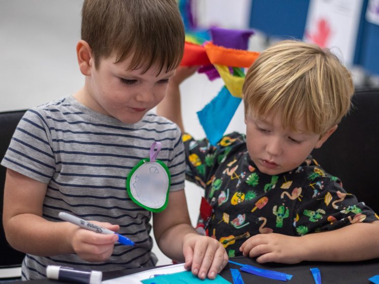 Image of Two young boys sit together at a table, collaborating on an art project. One holds a marker and draws on paper, whilst the other carefully places blue tissue pieces. Colourful supplies and drawings are visible around them.