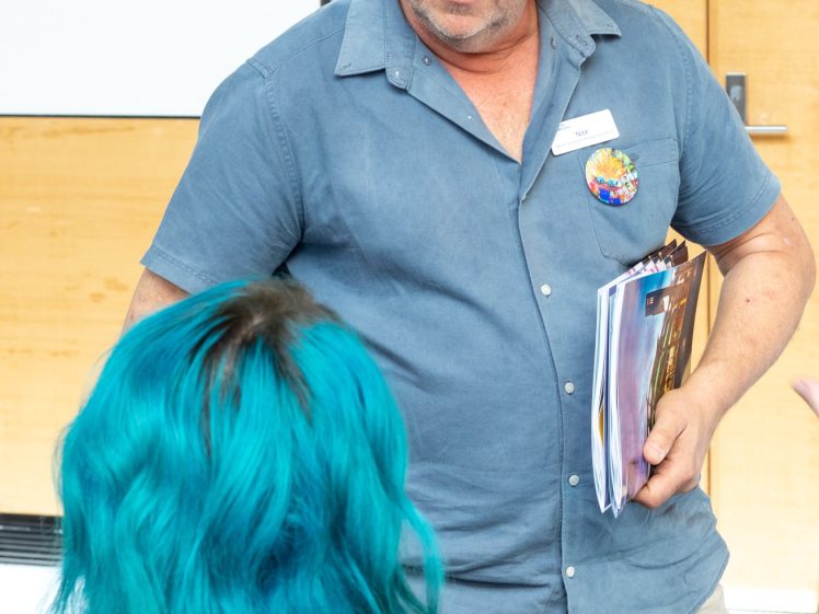 Image of A man in a blue shirt holds magazines and talks to a person with bright blue hair, seen from behind, in a brightly lit indoor setting with wooden walls and a framed picture on the wall.