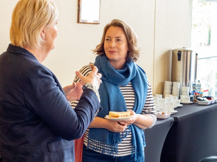 Image of Two women are talking at a coffee break. One woman holds a sandwich and wears a blue scarf and striped shirt; the other gestures with her hands. A refreshments table with coffee cups and a large coffee urn is in the background.