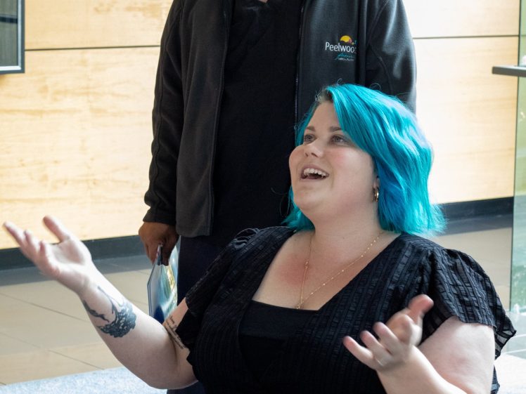 Image of A woman with bright blue hair sits in a wheelchair, smiling and gesturing with her hands. A man standing behind her is also smiling. They appear to be in a modern indoor setting with wooden walls.
