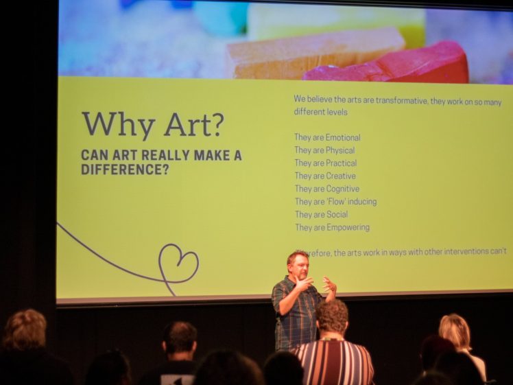 Image of A man stands on stage speaking to an audience in front of a large presentation slide titled Why Art? Can art really make a difference? with a list of ways the arts are transformative.