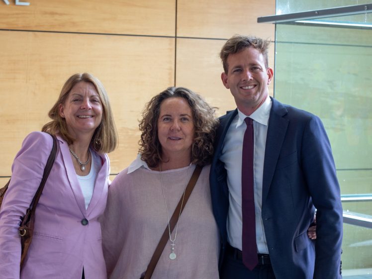 Image of Three people stand closely together and smile at the camera. Two women, one in a light pink blazer and the other in a light jumper, are next to a man in a dark suit and tie. They are indoors in a modern setting.