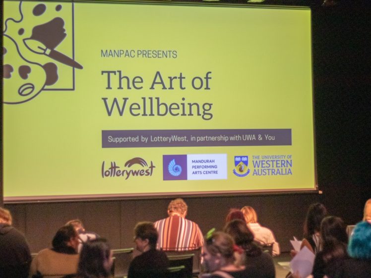 Image of A group of people sit facing a large screen displaying The Art of Wellbeing event, supported by Lotterywest, Mandurah Performing Arts Centre, and the University of Western Australia.