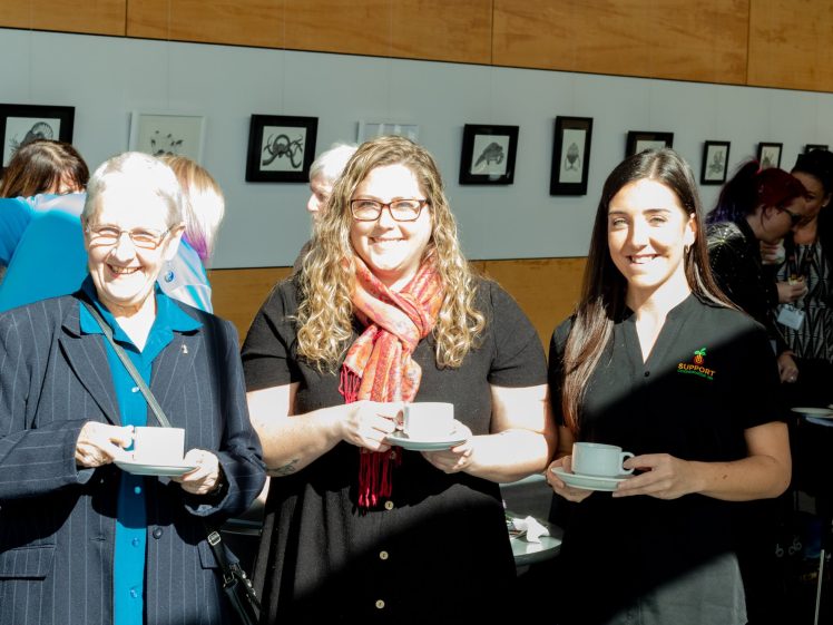 Image of Three women standing indoors, smiling and holding white cups and saucers. Artworks hang on the wall behind them, and other people are mingling in the background. Bright sunlight illuminates the scene.