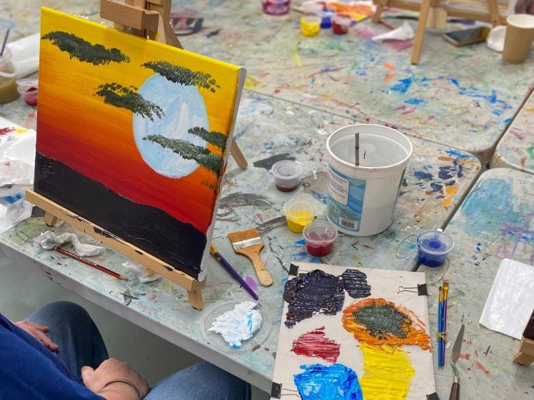 Image of A person sits at a messy art table, painting a sunset landscape with dark clouds and a bright sun on a canvas. Paints, brushes, and a palette with mixed colours are scattered around the workspace.