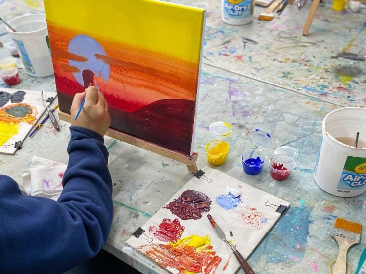 Image of A person in a blue jumper paints a sunset landscape on a canvas, using red, yellow, and blue paints. Art supplies, brushes, and a palette with mixed colours are spread across a messy table.
