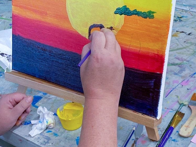 Image of A person paints a sunset scene on a canvas, featuring a large yellow sun, colourful sky, and dark water. Art supplies, brushes, and paint containers are scattered on the table around the easel.